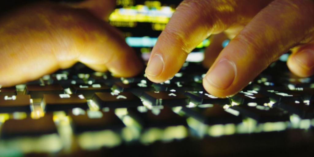 Mãos digitando rapidamente em um teclado iluminado por tela de computador.